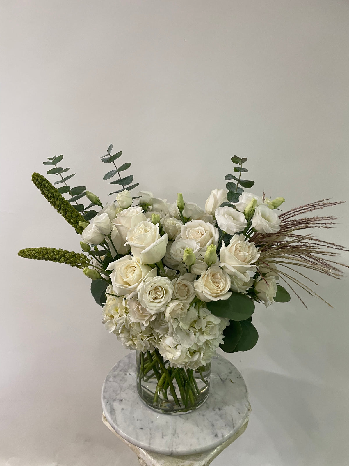 A floral arrangement of white roses, white hydrangeas, green eucalyptus stems, and dried grasses in a clear glass vase, displayed on a round white marble pedestal against a plain light gray wall.